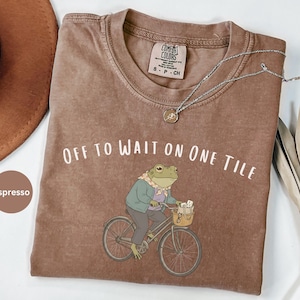 Op de afbeelding: Een espresso-kleurig t-shirt met een afbeelding van een kikker met een sjaal en jas op een fiets. De tekst "OFF TO WAIT ON ONE TILE" is in het wit boven de kikker gedrukt. Het t-shirt heeft een ronde hals en een Comfort Colors label.