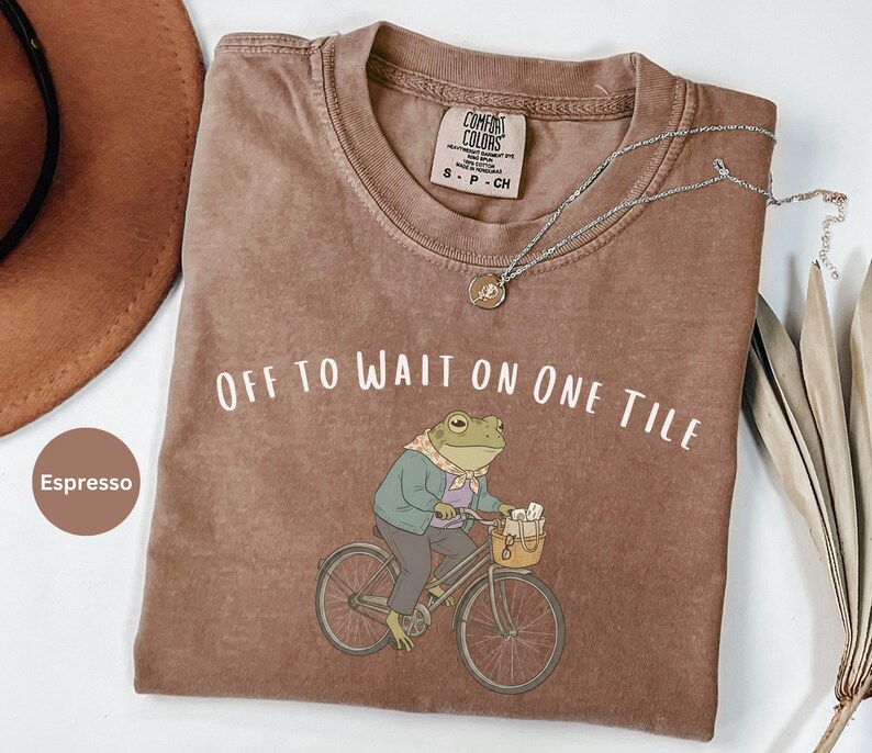 Op de afbeelding: Een espresso-kleurig t-shirt met een afbeelding van een kikker met een sjaal en jas op een fiets. De tekst "OFF TO WAIT ON ONE TILE" is in het wit boven de kikker gedrukt. Het t-shirt heeft een ronde hals en een Comfort Colors label.