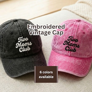 May include: Two embroidered vintage caps, one black and one pink, are displayed. Each cap features the text "Two Moms Club" in a contrasting color. The image also includes text indicating that six colors are available.