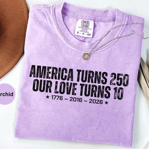 May include: A light purple short-sleeve t-shirt with a distressed black graphic print that reads AMERICA TURNS 250 OUR LOVE TURNS 10 1776 - 2016 - 2026 with stars. The shirt features a crew neckline and a white interior label with black text.