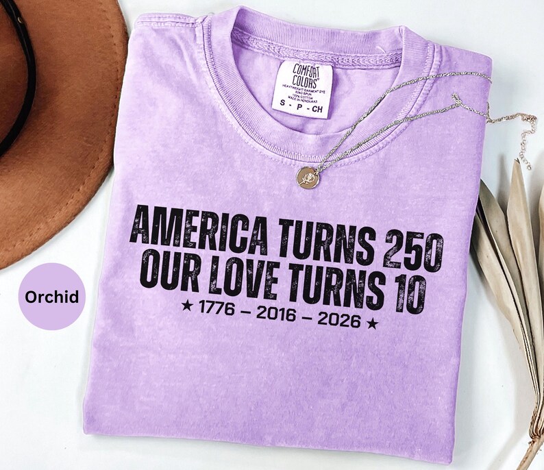 May include: A light purple short-sleeve t-shirt with a distressed black graphic print that reads AMERICA TURNS 250 OUR LOVE TURNS 10 1776 - 2016 - 2026 with stars. The shirt features a crew neckline and a white interior label with black text.
