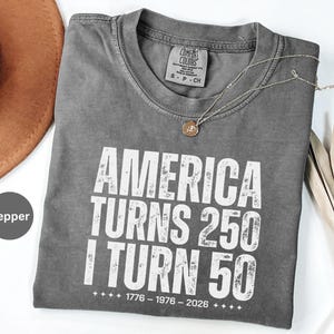 May include: Pepper-colored, short-sleeved t-shirt with the text "AMERICA TURNS 250 I TURN 50" in white. The shirt also has the dates "1776 - 1976 - 2026" printed below the text. A silver necklace with a pendant is on the shirt.