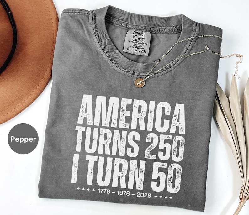 May include: Pepper-colored, short-sleeved t-shirt with the text "AMERICA TURNS 250 I TURN 50" in white. The shirt also has the dates "1776 - 1976 - 2026" printed below the text. A silver necklace with a pendant is on the shirt.
