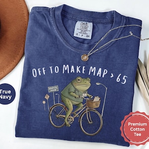 May include: A true navy blue cotton t-shirt featuring a graphic of a green frog wearing a medical scrub top and riding a bicycle. The text reads OFF TO MAKE MAP > 65. The frog has a basket on the handlebars containing a patient chart and a stethoscope.