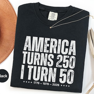 May include: Black t-shirt with white distressed text that reads "AMERICA TURNS 250 I TURN 50" with the years 1776-1976-2026 below. A gold necklace with a star pendant rests on the shirt. A brown hat is visible in the background.