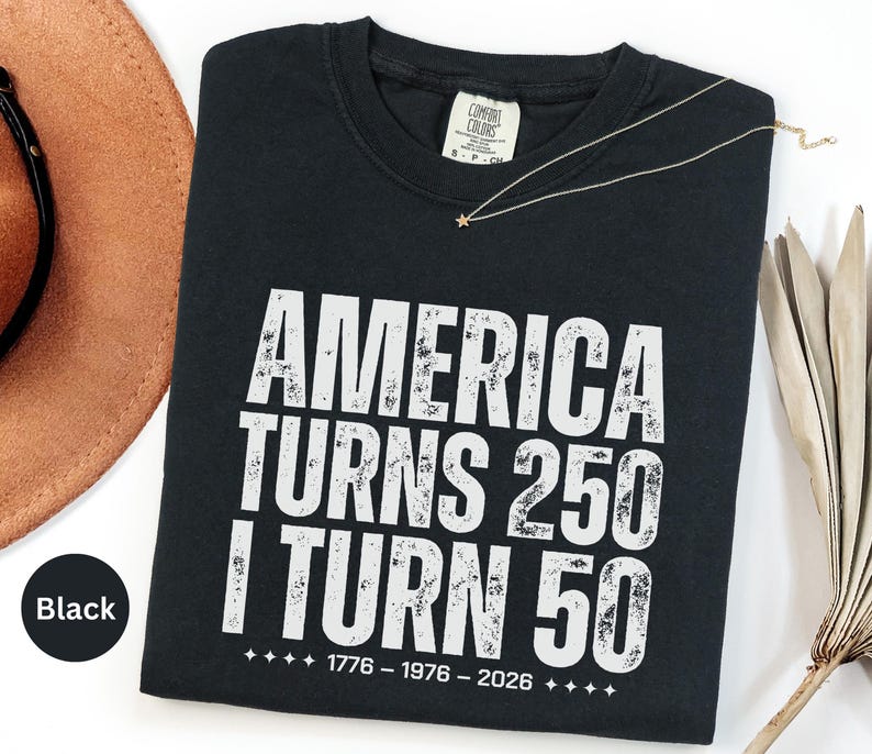 May include: Black t-shirt with white distressed text that reads "AMERICA TURNS 250 I TURN 50" with the years 1776-1976-2026 below. A gold necklace with a star pendant rests on the shirt. A brown hat is visible in the background.