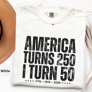 May include: White t-shirt with the text "AMERICA TURNS 250 I TURN 50" in distressed black lettering. The shirt includes the years 1776-1976-2026. A gold necklace with a star pendant and a brown hat are also visible.