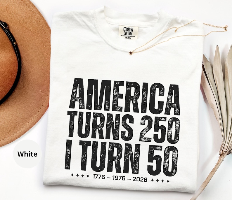 May include: White t-shirt with the text "AMERICA TURNS 250 I TURN 50" in distressed black lettering. The shirt includes the years 1776-1976-2026. A gold necklace with a star pendant and a brown hat are also visible.