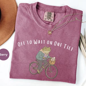 Op de afbeelding: Een bessenkleurig t-shirt met korte mouwen en een afbeelding van een kikker op een fiets met een mandje. De tekst "OFF TO WAIT ON ONE TILE" staat boven de kikker gedrukt. Het shirt heeft een Comfort Colors label met maat S-P-CH.