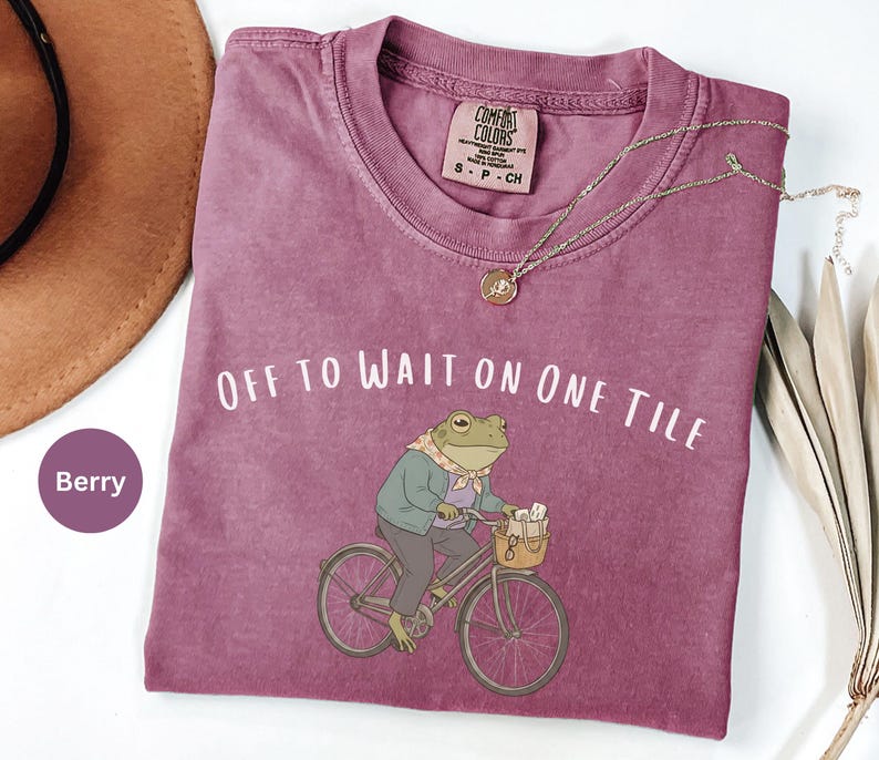 Op de afbeelding: Een bessenkleurig t-shirt met korte mouwen en een afbeelding van een kikker op een fiets met een mandje. De tekst "OFF TO WAIT ON ONE TILE" staat boven de kikker gedrukt. Het shirt heeft een Comfort Colors label met maat S-P-CH.