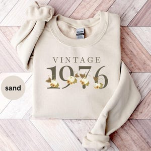 May include: A sand-colored crewneck sweatshirt featuring the text VINTAGE 1976 in dark lettering. The numbers are decorated with illustrations of small white pumpkins, autumn leaves, and vines. The garment has long sleeves and a ribbed neckline.