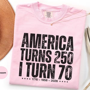May include: A light pink t-shirt with the text "AMERICA TURNS 250 I TURN 70" in black, along with the years 1776-1956-2026. The shirt is folded and has a tag at the neckline.