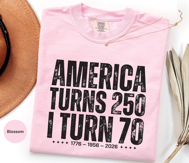 May include: A light pink t-shirt with the text "AMERICA TURNS 250 I TURN 70" in black, along with the years 1776-1956-2026. The shirt is folded and has a tag at the neckline.