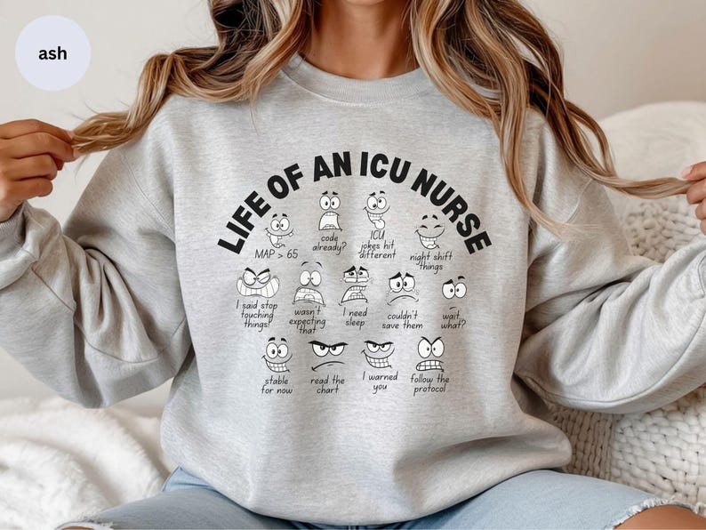 May include: A light gray sweatshirt with the text "LIFE OF AN ICU NURSE" in black. Below the text are various cartoon faces with different expressions and phrases like "code already?" and "night shift things".