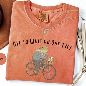 Op de afbeelding: Een yam-kleurig T-shirt met korte mouwen en een afbeelding van een kikker in een jas en sjaal op een fiets. De tekst "OFF TO WAIT ON ONE TILE" staat in het zwart boven de kikker gedrukt. Het shirt heeft een ronde hals en een label.