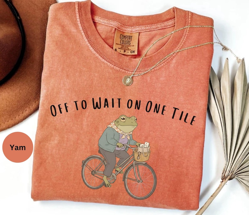 Op de afbeelding: Een yam-kleurig T-shirt met korte mouwen en een afbeelding van een kikker in een jas en sjaal op een fiets. De tekst "OFF TO WAIT ON ONE TILE" staat in het zwart boven de kikker gedrukt. Het shirt heeft een ronde hals en een label.