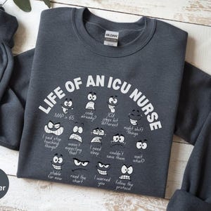 May include: Dark heather gray sweatshirt with the text "LIFE OF AN ICU NURSE" in white. The design includes various cartoon faces with phrases like "MAP > 65" and "night shift things".