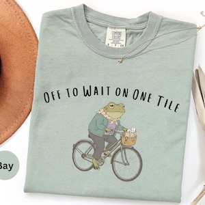Op de afbeelding: Een saliegroen t-shirt met een afbeelding van een kikker in een jas en sjaal op een fiets. De tekst boven de kikker luidt "OFF TO WAIT ON ONE TILE". Het t-shirt heeft een ronde hals en een Comfort Colors merklabel bij de kraag.