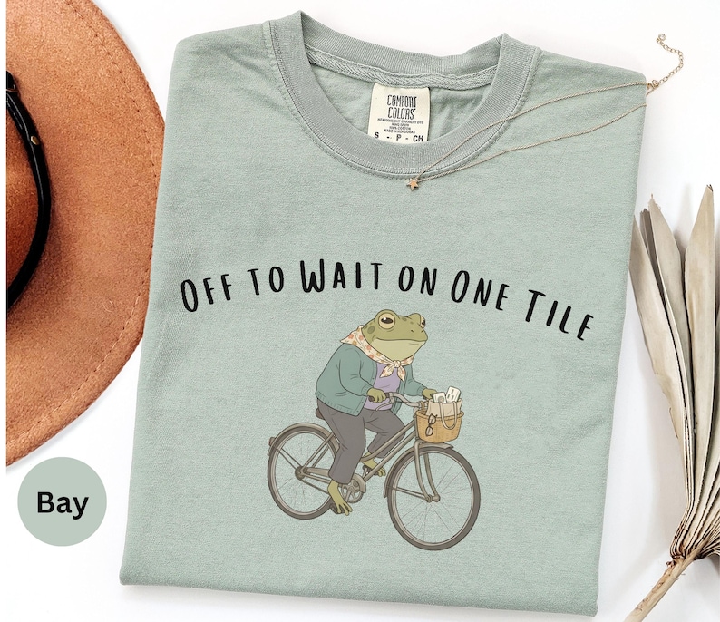 Op de afbeelding: Een saliegroen t-shirt met een afbeelding van een kikker in een jas en sjaal op een fiets. De tekst boven de kikker luidt "OFF TO WAIT ON ONE TILE". Het t-shirt heeft een ronde hals en een Comfort Colors merklabel bij de kraag.