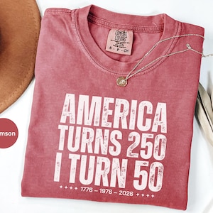 May include: Crimson-colored t-shirt with the text "AMERICA TURNS 250 I TURN 50" in white, with the years 1776-1976-2026 below. A gold necklace with a pendant rests on the shirt. A brown hat is partially visible.