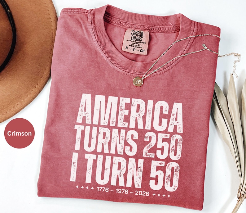 May include: Crimson-colored t-shirt with the text "AMERICA TURNS 250 I TURN 50" in white, with the years 1776-1976-2026 below. A gold necklace with a pendant rests on the shirt. A brown hat is partially visible.