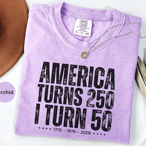 May include: Orchid-colored t-shirt with the text "AMERICA TURNS 250 I TURN 50" in black. The shirt has a crew neck and short sleeves. A silver necklace with a pendant is draped across the shirt.