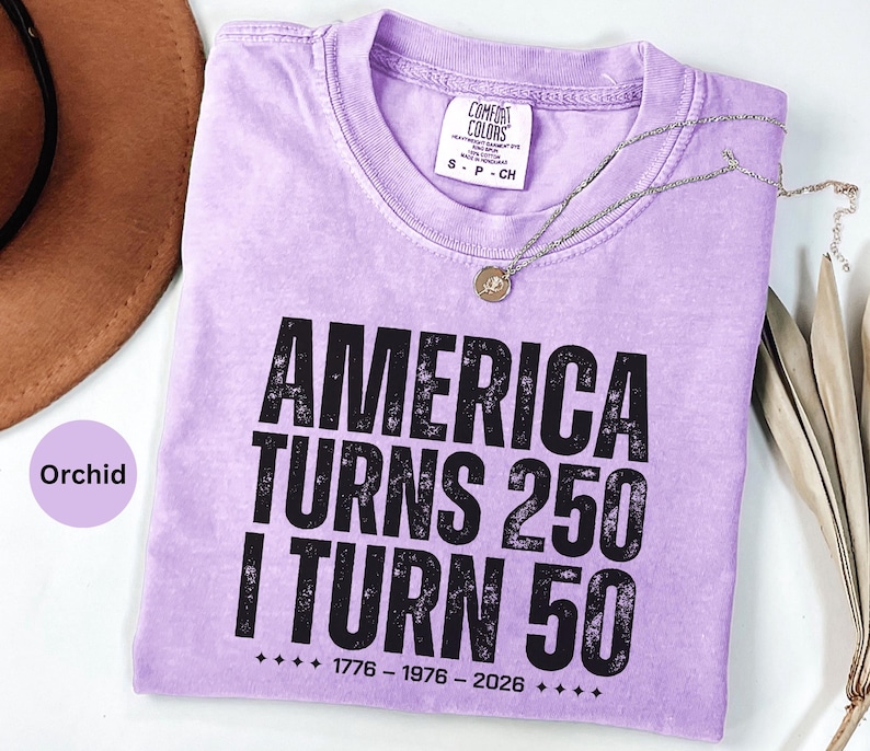 May include: Orchid-colored t-shirt with the text "AMERICA TURNS 250 I TURN 50" in black. The shirt has a crew neck and short sleeves. A silver necklace with a pendant is draped across the shirt.