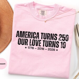 May include: A light pink short-sleeve t-shirt with black text that reads AMERICA TURNS 250 OUR LOVE TURNS 10, followed by the years 1776 - 2016 - 2026 with a star on each side. The shirt features a crew neckline and a Comfort Colors brand label.