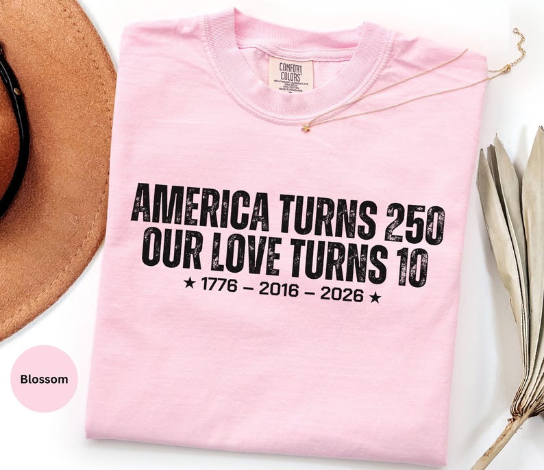 May include: A light pink short-sleeve t-shirt with black text that reads AMERICA TURNS 250 OUR LOVE TURNS 10, followed by the years 1776 - 2016 - 2026 with a star on each side. The shirt features a crew neckline and a Comfort Colors brand label.