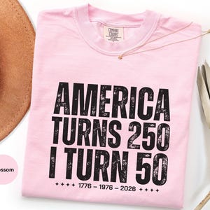 May include: A light pink t-shirt with the text "AMERICA TURNS 250 I TURN 50" in black, distressed font. The shirt also features the years 1776-1976-2026. A gold necklace is draped across the shirt.