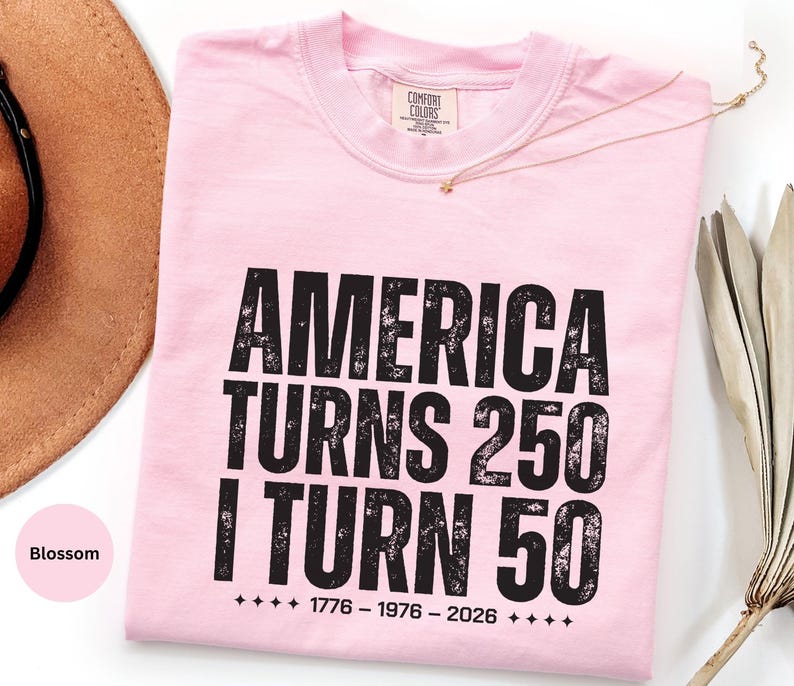May include: A light pink t-shirt with the text "AMERICA TURNS 250 I TURN 50" in black, distressed font. The shirt also features the years 1776-1976-2026. A gold necklace is draped across the shirt.