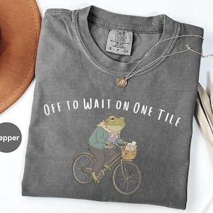 Op de afbeelding: Een peperkleurig t-shirt met een afbeelding van een kikker in een jas en sjaal op een fiets met een mandje. De tekst "OFF TO WAIT ON ONE TILE" staat in het wit boven de kikker gedrukt. Het t-shirt heeft een ronde hals en een label in de nek.