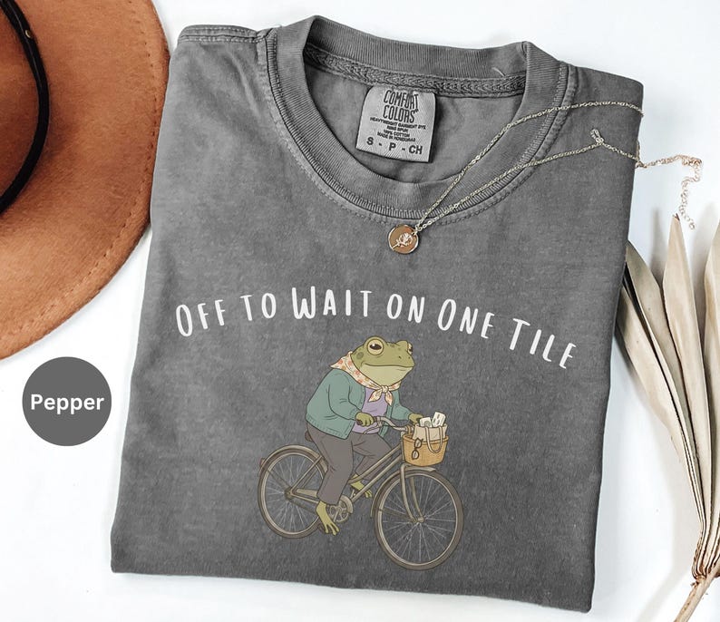 Op de afbeelding: Een peperkleurig t-shirt met een afbeelding van een kikker in een jas en sjaal op een fiets met een mandje. De tekst "OFF TO WAIT ON ONE TILE" staat in het wit boven de kikker gedrukt. Het t-shirt heeft een ronde hals en een label in de nek.