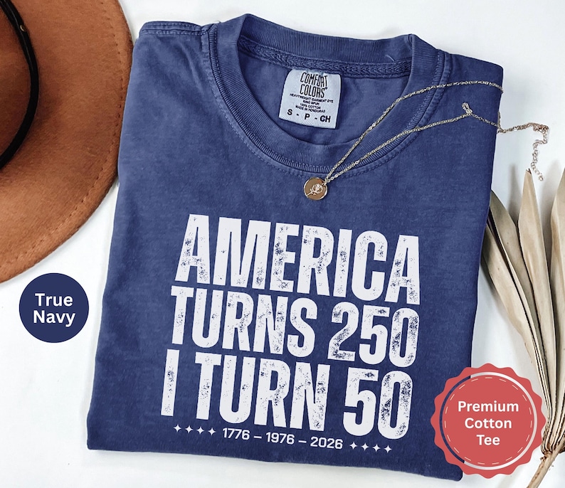 May include: A navy blue t-shirt with the text "AMERICA TURNS 250 I TURN 50" in white. The shirt has a crew neck and short sleeves. A gold necklace with a pendant is draped on the shirt. The shirt is labeled as a "Premium Cotton Tee".