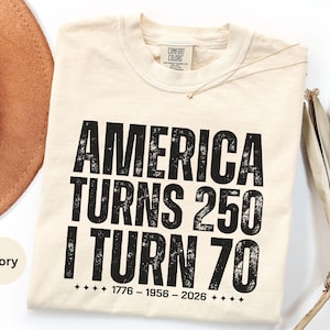 May include: Ivory-colored t-shirt with the text "AMERICA TURNS 250 I TURN 70" in bold, distressed black font. The shirt includes the dates 1776-1956-2026. A brown hat and dried palm leaf are partially visible in the background.