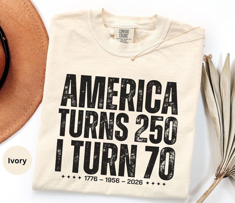 May include: Ivory-colored t-shirt with the text "AMERICA TURNS 250 I TURN 70" in bold, distressed black font. The shirt includes the dates 1776-1956-2026. A brown hat and dried palm leaf are partially visible in the background.