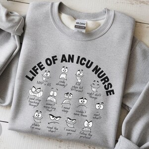 May include: Sport grey sweatshirt with the text "LIFE OF AN ICU NURSE" in black, with various cartoon faces and phrases. The sweatshirt is made of a soft material and has a crew neck.
