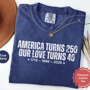 May include: A navy blue short-sleeve t-shirt with a distressed white graphic print that reads: AMERICA TURNS 250 OUR LOVE TURNS 40, followed by the years 1776 - 1986 - 2026 with stars on each side. The shirt features a Comfort Colors brand neck label.