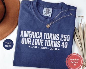 May include: A navy blue short-sleeve t-shirt with a distressed white graphic print that reads: AMERICA TURNS 250 OUR LOVE TURNS 40, followed by the years 1776 - 1986 - 2026 with stars on each side. The shirt features a Comfort Colors brand neck label.