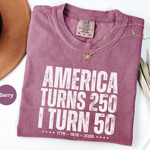 May include: Berry-colored t-shirt with the text "America Turns 250, I Turn 50" in white. The shirt has a crew neck and short sleeves. A gold necklace with a pendant is draped across the shirt.