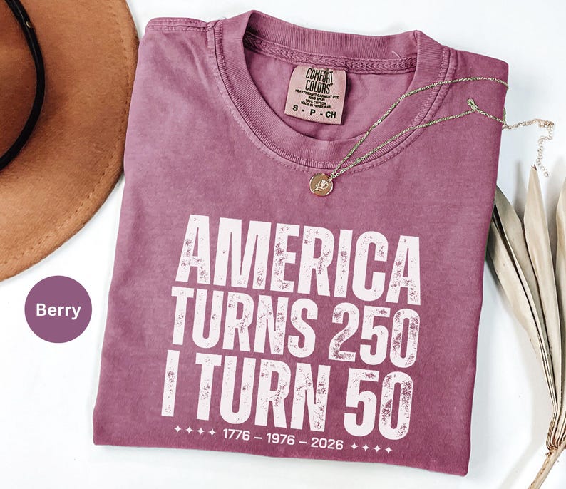 May include: Berry-colored t-shirt with the text "America Turns 250, I Turn 50" in white. The shirt has a crew neck and short sleeves. A gold necklace with a pendant is draped across the shirt.