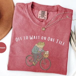 Op de afbeelding: Een karmozijnrood t-shirt met ronde hals en een afbeelding van een kikker op een fiets met mandje, gekleed in een jasje en sjaal. De tekst luidt OFF TO WAIT ON ONE TILE. Het shirt heeft een Comfort Colors label en een kleine hanger.