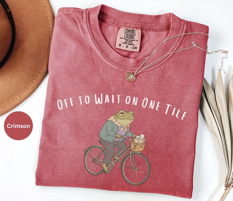Op de afbeelding: Een karmozijnrood t-shirt met ronde hals en een afbeelding van een kikker op een fiets met mandje, gekleed in een jasje en sjaal. De tekst luidt OFF TO WAIT ON ONE TILE. Het shirt heeft een Comfort Colors label en een kleine hanger.