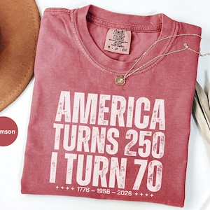 May include: Crimson-colored t-shirt with the text "AMERICA TURNS 250 I TURN 70" in white block letters. The shirt includes the years 1776-1956-2026. A gold necklace with a pendant rests on the shirt. The shirt is part of the Comfort Colors brand.