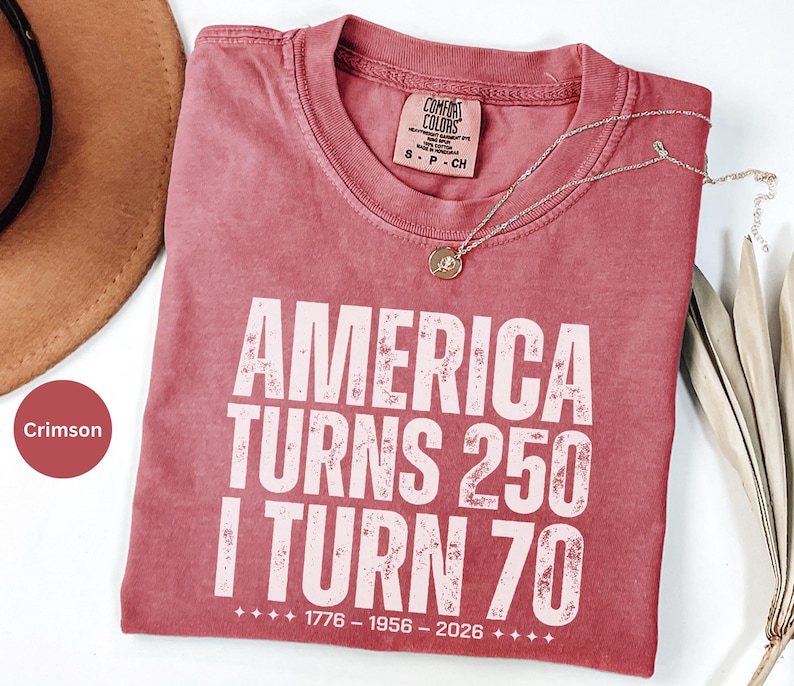 May include: Crimson-colored t-shirt with the text "AMERICA TURNS 250 I TURN 70" in white block letters. The shirt includes the years 1776-1956-2026. A gold necklace with a pendant rests on the shirt. The shirt is part of the Comfort Colors brand.