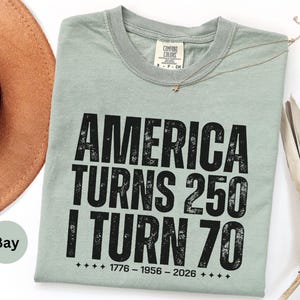May include: A sage green t-shirt with the text "AMERICA TURNS 250 I TURN 70" in black. The shirt also has the dates 1776-1956-2026. A brown hat is partially visible in the upper left corner.