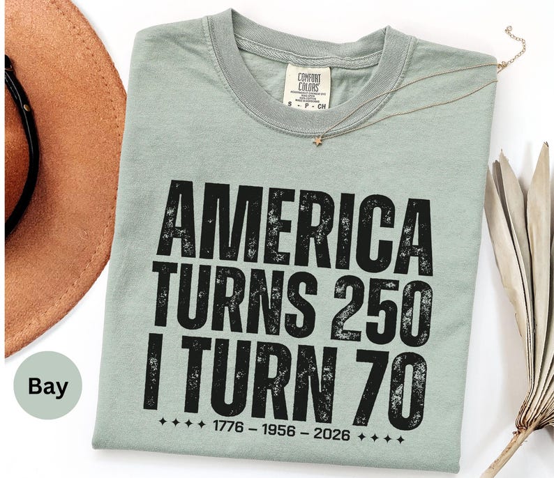May include: A sage green t-shirt with the text "AMERICA TURNS 250 I TURN 70" in black. The shirt also has the dates 1776-1956-2026. A brown hat is partially visible in the upper left corner.