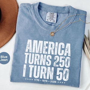 May include: Blue jean colored t-shirt with the text "AMERICA TURNS 250 I TURN 50" in white. The shirt has a crew neck and a small tag on the collar. A gold necklace with a pendant is draped across the shirt.