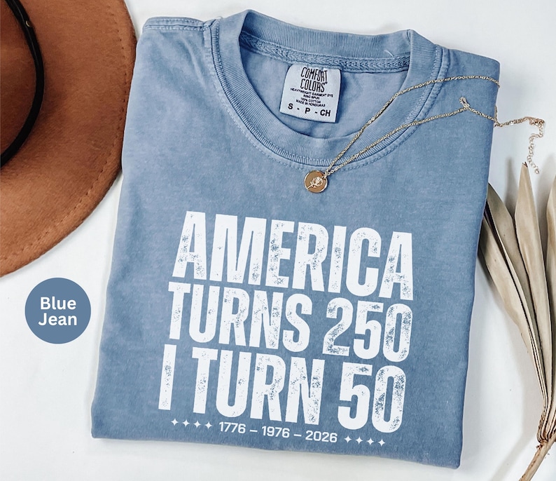 May include: Blue jean colored t-shirt with the text "AMERICA TURNS 250 I TURN 50" in white. The shirt has a crew neck and a small tag on the collar. A gold necklace with a pendant is draped across the shirt.