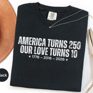 May include: A black short-sleeved t-shirt featuring white distressed text that reads AMERICA TURNS 250 OUR LOVE TURNS 10, with the years 1776 - 2016 - 2026 and two stars printed below. The shirt has a Comfort Colors brand label at the neck.